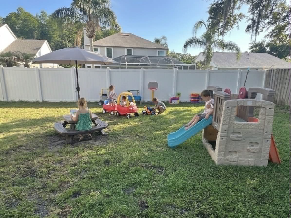 Children playing together in the fenced backyard at Amany's Home Daycare, with a playhouse slide, ride-on toys, and picnic table
